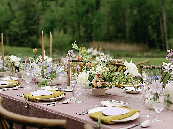 Reception tablescape with an outdoor reception table set with floral centerpiece, bud vases and taper candles on a lawn among trees and tables