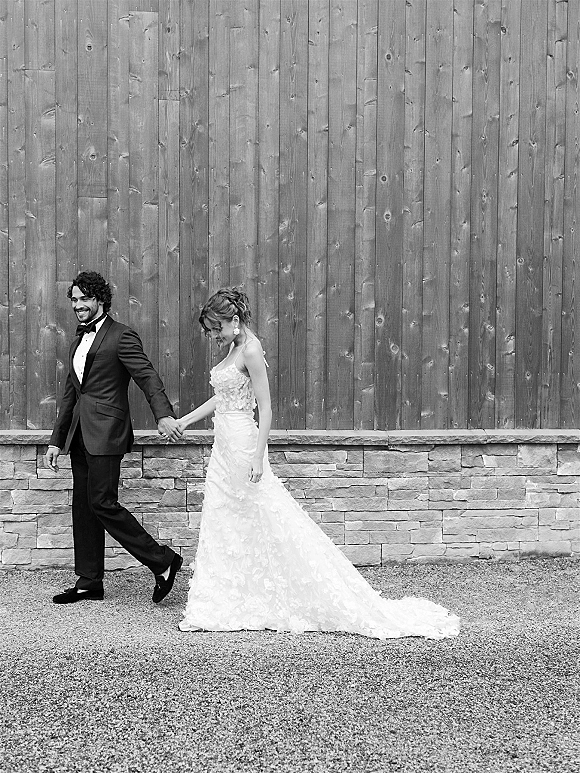 Couple portrait of bride and groom holding hands, groom in tuxedo and bride in lace dress with updo, beside rustic wood and stone wall
