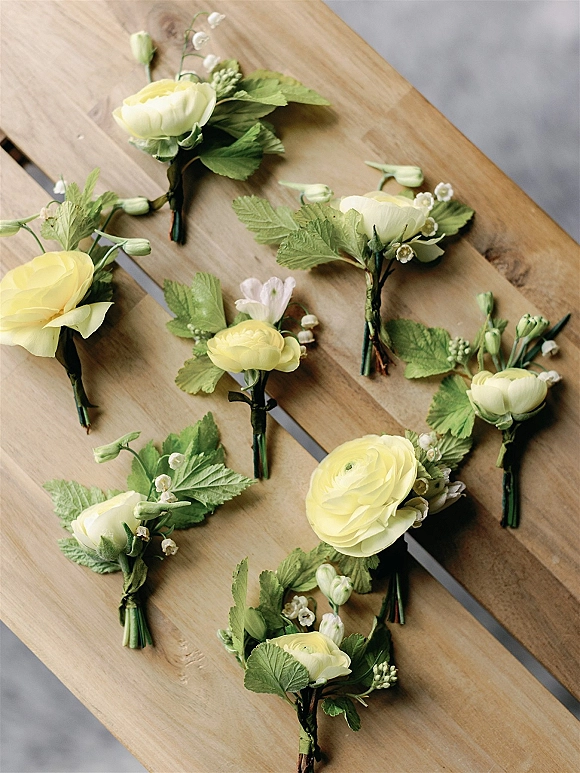 Wedding boutonnieres arranged in a flat lay on a wood table, with yellow flowers and greenery tied with floral tape over concrete floor