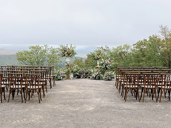 Ceremony setup for an outdoor wedding ceremony with an aisle of wood chairs, floral pedestal urns, and mountain view terrace backdrop