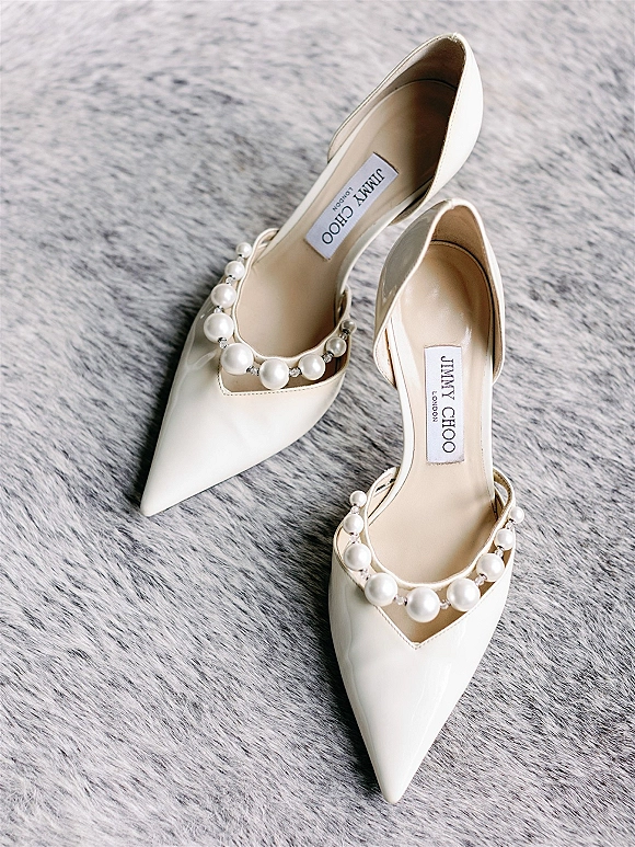 Wedding shoes with pearl strap in ivory patent leather, pointed toe heels displayed on a gray fur rug with designer label visible