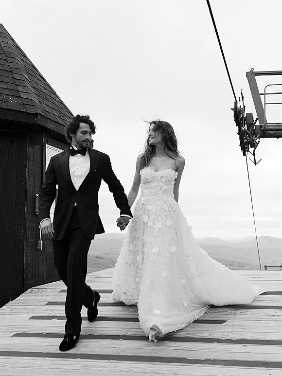 Couple portrait of bride and groom holding hands, walking on a wood deck by a building, her gown train trailing toward distant hills.