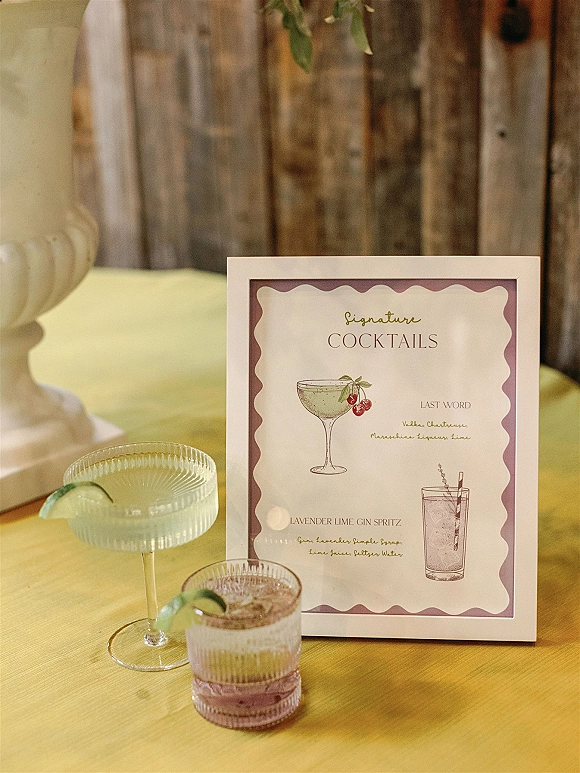 Signature cocktail sign with illustrated wedding signature cocktails in a framed menu, showing coupe and rocks glasses with lime on a wood table