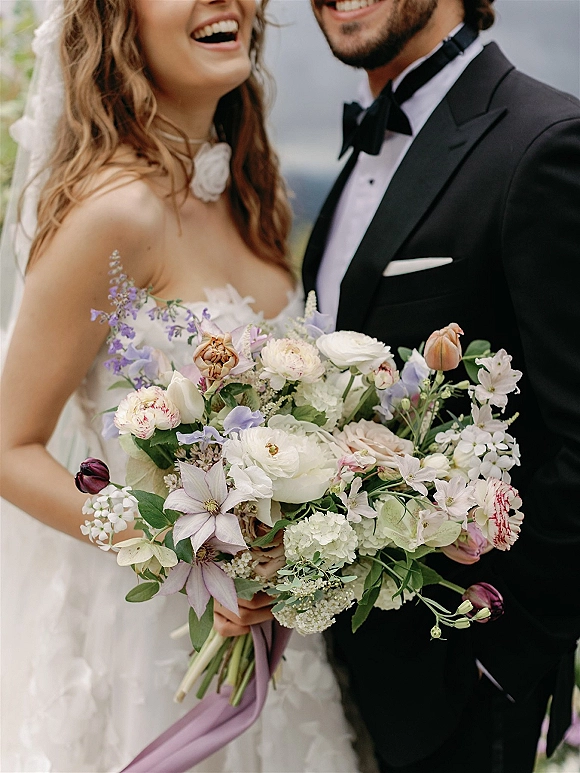 Couple portrait of bride and groom laughing close up, her veil and strapless dress beside his tuxedo, holding a bouquet with ribbon tails outdoors