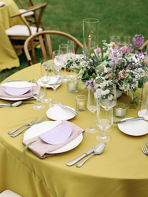 Reception tablescape with a mustard wedding tablecloth, pastel floral centerpiece, taper candles, and blush napkins on a grass lawn outdoors