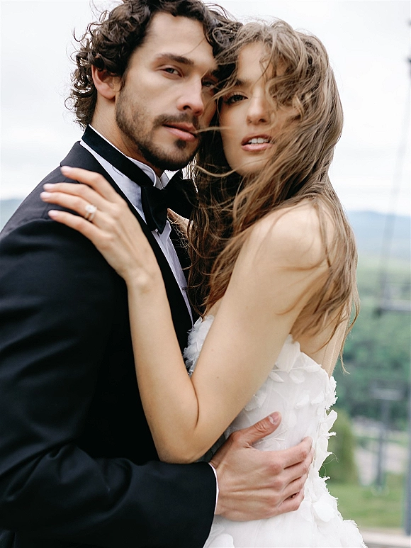 Couple portrait of bride and groom close up, her hand on his shoulder showing an engagement ring against distant hills and sky