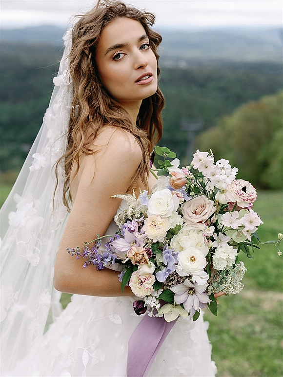 Bridal portrait of a bride holding bouquet with a lace veil and pastel blooms, turned in profile against misty mountain hills