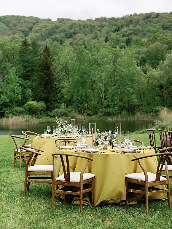Reception tablescape with mustard tablecloths, floral centerpieces, taper candles, and place settings on round tables by a pond and trees