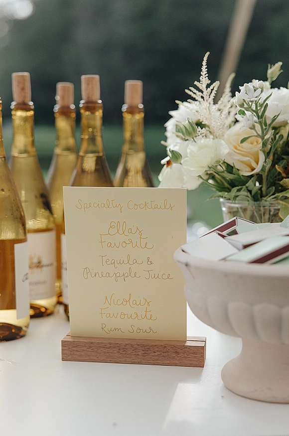 Wedding bar sign with specialty cocktails listed in calligraphy on a wooden holder beside wine bottles and rose greenery on an outdoor table