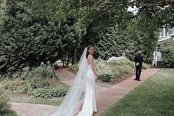 First look moment as bride approaches groom on a brick garden path, her long cathedral veil trailing behind in soft natural light