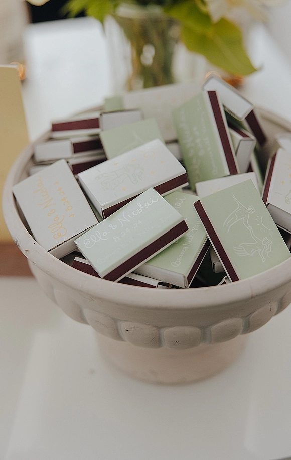 Wedding matchboxes piled in a ceramic bowl, personalized wedding favor design on a white tabletop with blurred greenery behind