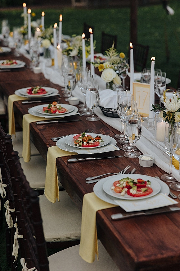 Reception tablescape on a long banquet table with white runner, taper and votive candles, florals, place cards, and glassware under a lawn tent