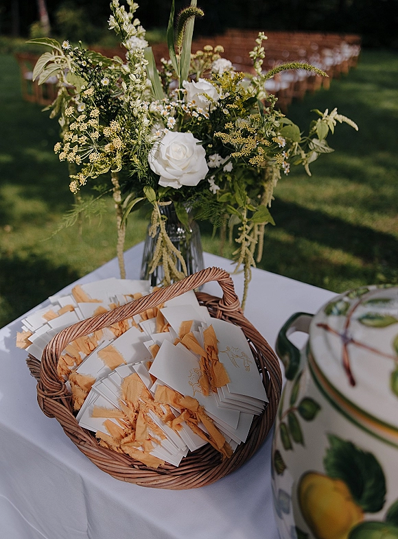 Wedding favors display with wicker basket and favor tags, tied with yellow ribbon beside white roses and wildflowers on a lawn table