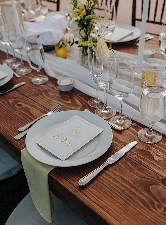 Reception tablescape with rustic wedding table setting on a farmhouse wood table, white runner, mixed glassware, taper candles, bud vase florals on patio