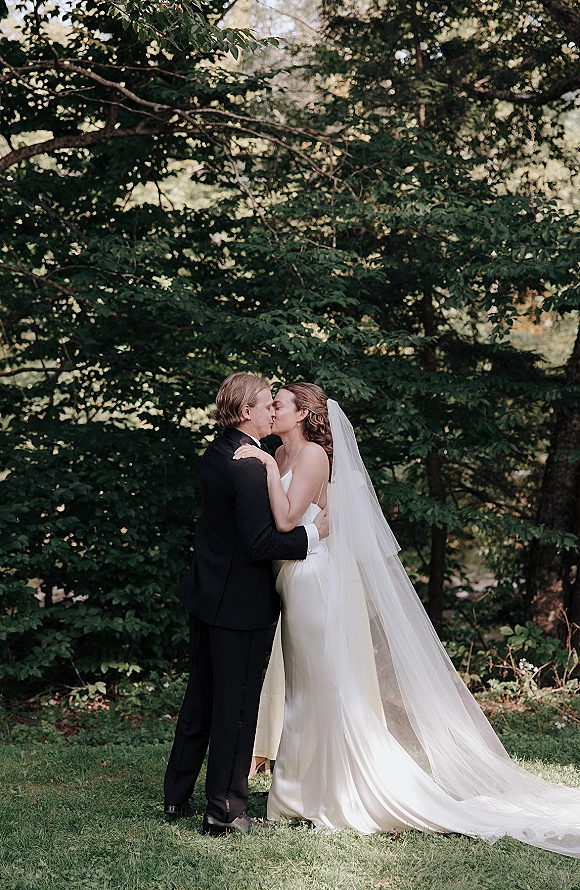 Wedding kiss portrait of bride and groom kissing on a garden lawn, her long cathedral veil trailing behind a strapless gown