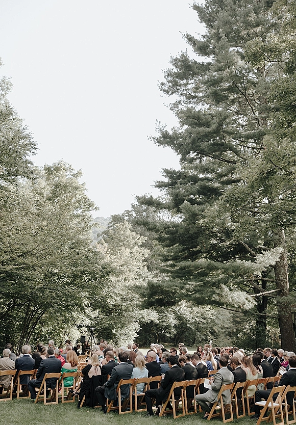 Outdoor wedding ceremony with wedding guests seated in rows on a grass lawn, wood folding chairs lined up under forest trees and open sky