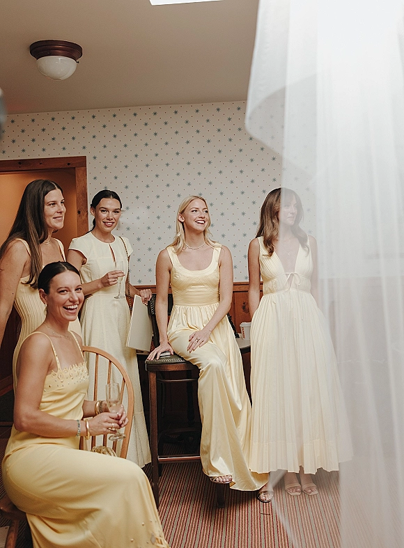 Bridesmaids portrait in yellow bridesmaid dresses holding champagne flutes, posed by a sheer curtain in a hotel room with patterned wallpaper