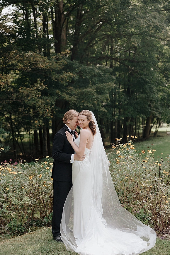 Couple portrait of bride and groom embrace, her cathedral veil draping on grass among wildflowers in a wooded park setting