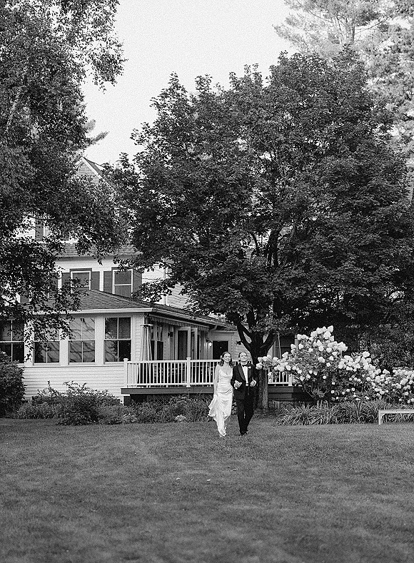 Couple portrait in black and white wedding portrait style, bride in satin dress holding hem beside groom in tux on estate lawn by porch