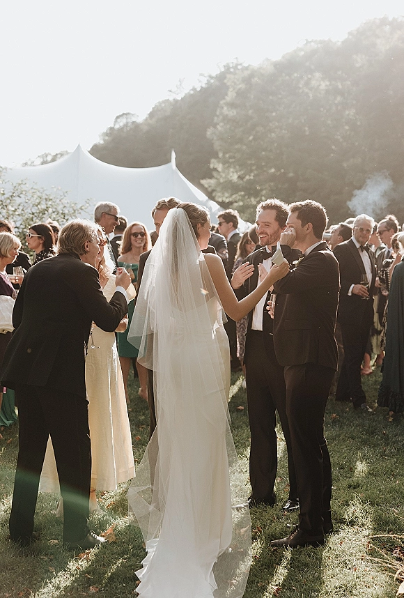 Wedding cocktail hour with guests mingling as the bride in a long veil and groom in tuxedos greet friends under a tent canopy on a sunny lawn