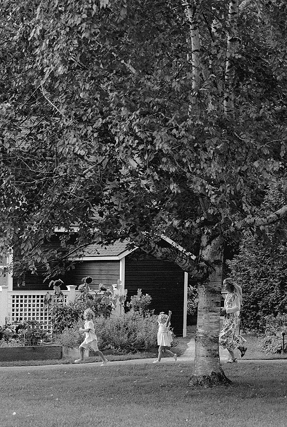 Wedding guest moments with kids at wedding as a woman in a floral maxi dress walks with children across a shady garden lawn by a house