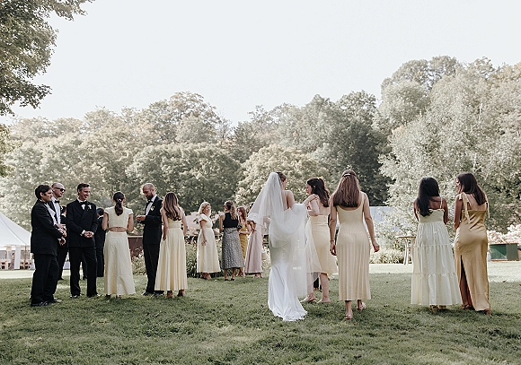 Wedding cocktail hour with guests mingling as the bride in veil stands with bridesmaids and tuxedoed groomsmen on a garden lawn near a tent