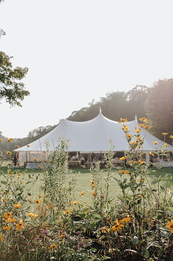Wedding reception tent in white sailcloth with string lights glowing above wildflowers on a sunny lawn with trees and blue sky