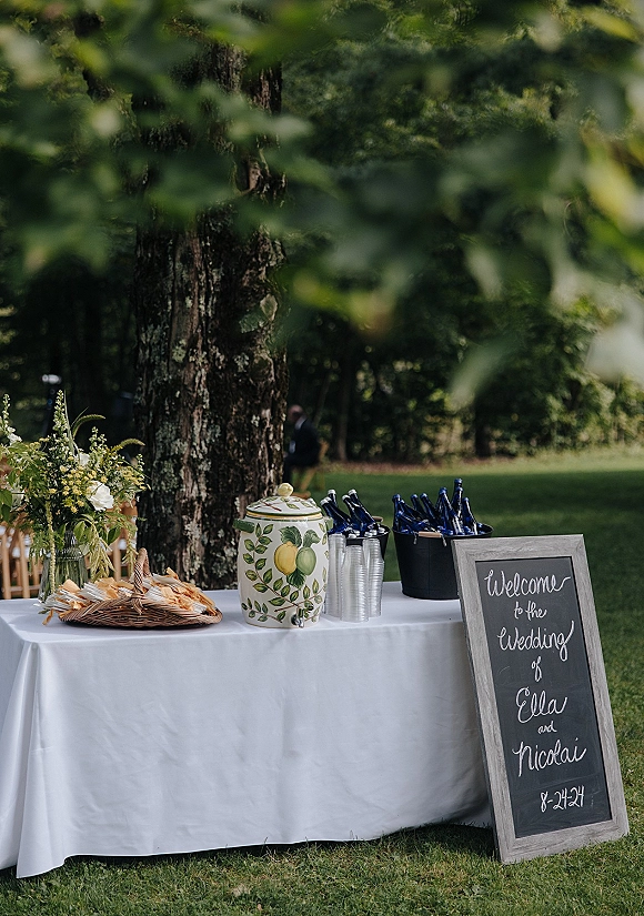 Wedding welcome table with a chalkboard welcome sign, white tablecloth, floral arrangement, and self-serve drinks set on a garden lawn