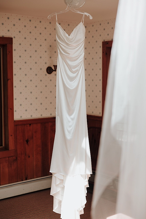 Wedding dress in satin hanging from a clear hanger, spaghetti straps and cowl neckline glowing in natural light by patterned wallpaper