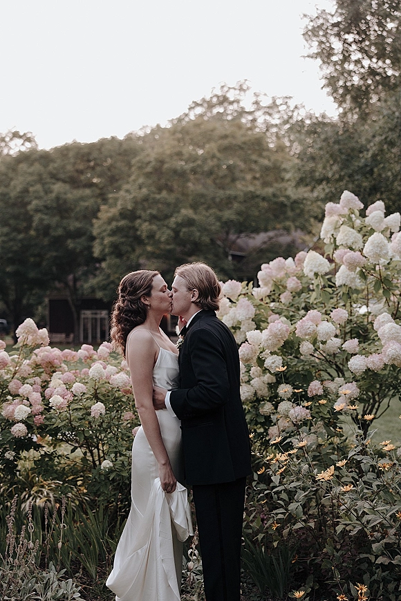 Wedding kiss portrait of bride and groom kissing in a lush garden, bride in a silk slip dress and groom in a black tuxedo boutonniere