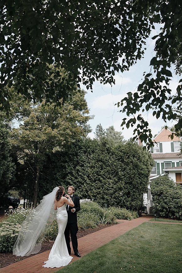 Couple portrait outdoors with bride in strapless wedding dress and long veil adjusting groom’s tie on a brick walkway in a garden