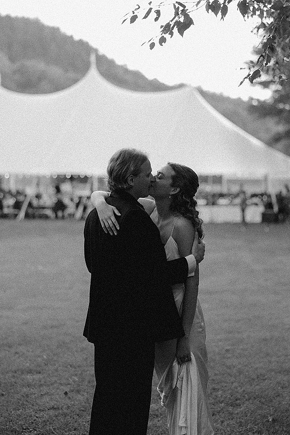 Wedding kiss portrait of bride and groom kissing under a white sailcloth tent, her satin dress and earrings against lawn, trees, hills
