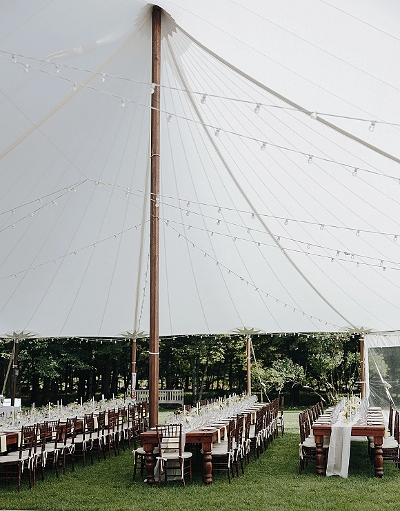 Reception tablescape under a sailcloth tent reception with string lights, long wooden farm tables, white runners, glassware, and taper candles on a garden lawn
