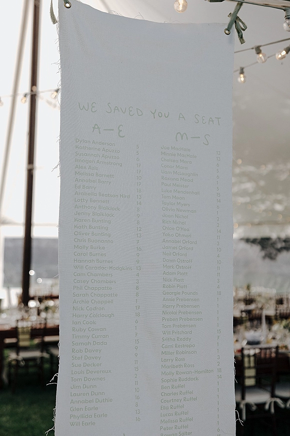 Wedding seating chart fabric banner with grommets and ribbon ties hanging under string lights in an outdoor tent reception setting