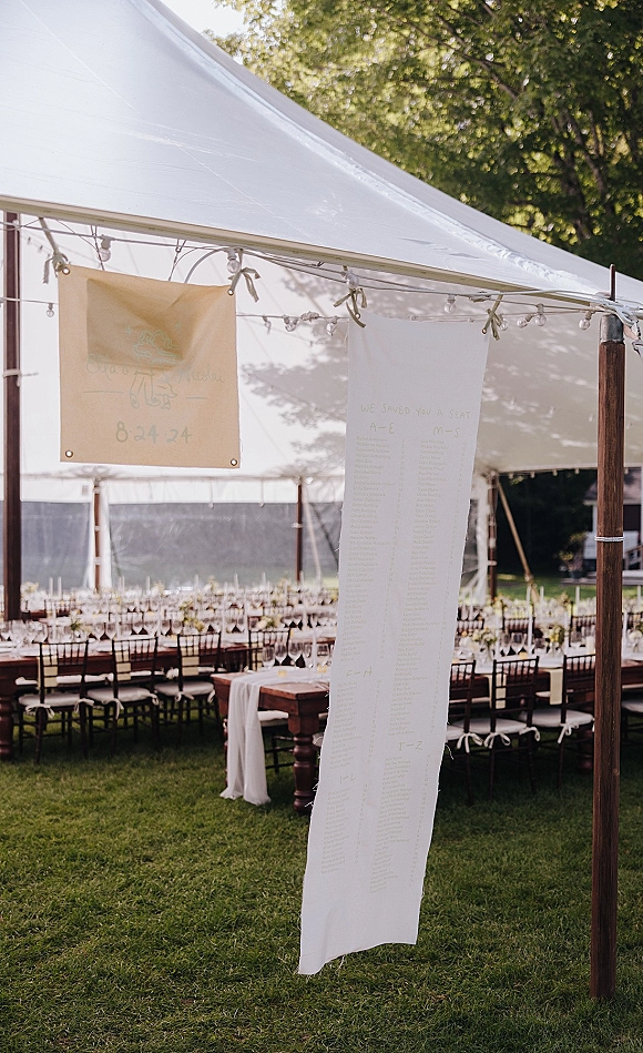 Seating chart display on a fabric seating chart, hanging from wooden poles with ribbon ties and string lights under a sailcloth tent.