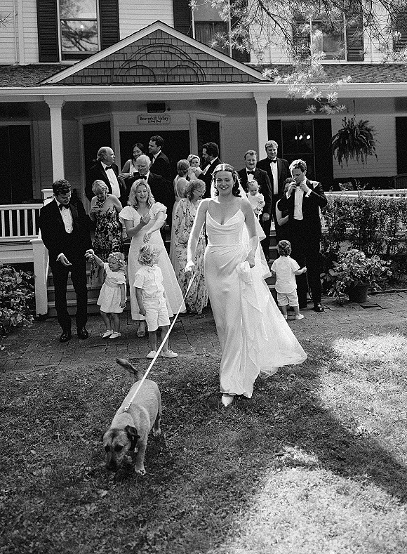 Wedding party photo in black and white of a veiled bride walking a dog on leash down front steps, surrounded by tuxedoed guests on a porch