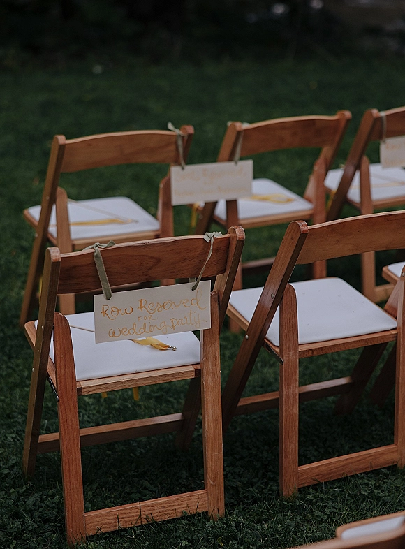 Ceremony seating with wedding reserved seating signs on wood folding chairs with white cushions, ribbon ties, and gold pen on a grass lawn