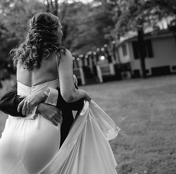 Couple portrait of groom holding bride in a lifted embrace, her backless dress and long train flowing under string lights on a lawn