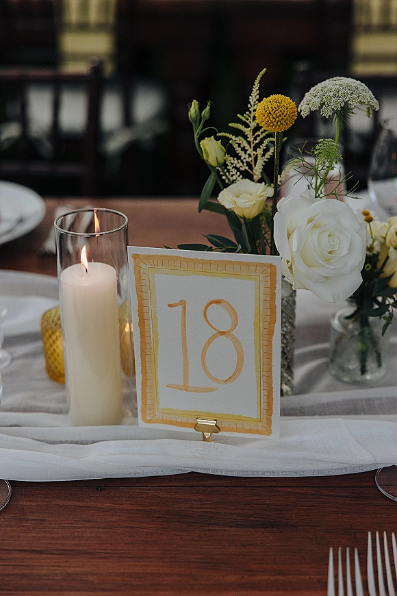 Wedding table number card in a gold holder beside a pillar candle in a glass hurricane, with white roses and yellow billy balls on wood table