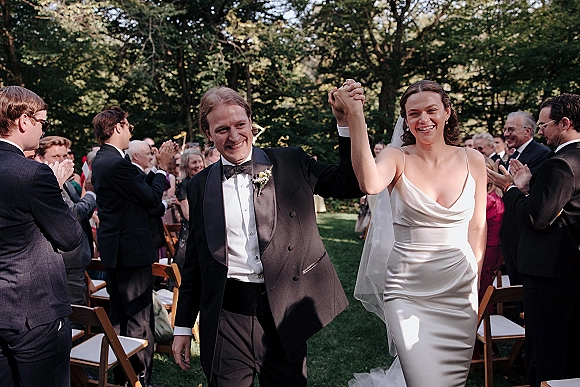 Wedding recessional as bride and groom walk hand in hand down the aisle, bride’s veil flowing in a sunlit garden with applauding guests