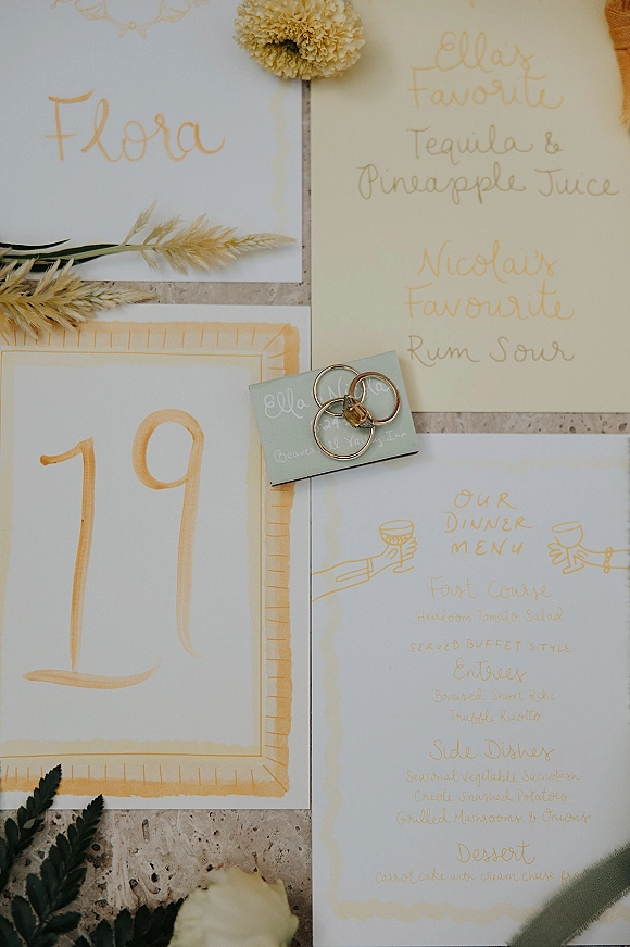 Wedding stationery flatlay with wedding menu card, calligraphy place cards and rings styled with dried wheat on a stone surface