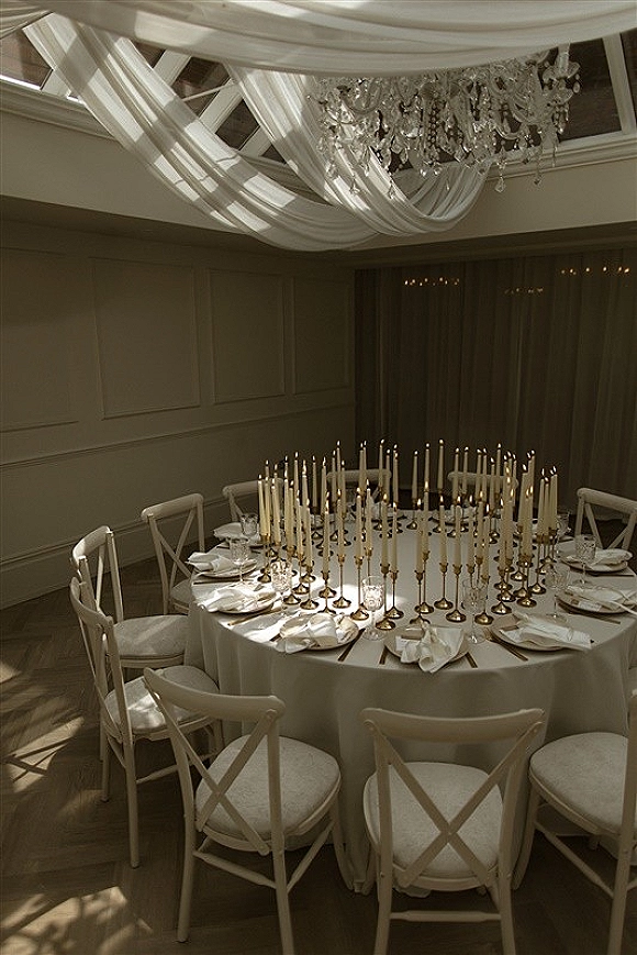 Reception tablescape with a round wedding reception table in white linens, taper candles in gold holders, and place settings beneath a chandelier and draped ceiling.