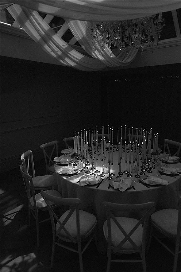 Reception tablescape with a candlelit wedding table of tall taper candles, white linens, chargers and glassware under chandelier and draped ceiling