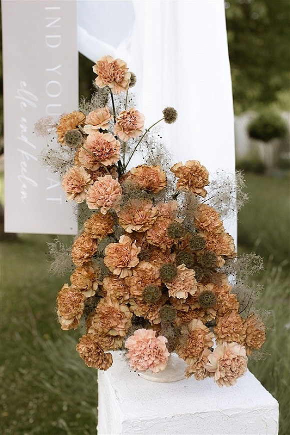 Wedding welcome sign on clear acrylic with peach carnations and baby’s breath, white fabric draping, set on an outdoor lawn with greenery