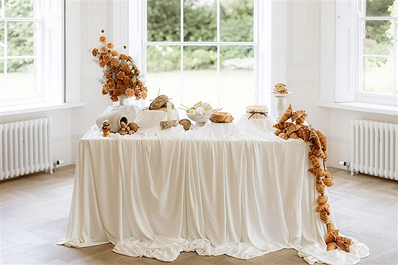 Wedding dessert table with bread and pastry table croissants, cookies, fruit and dried florals on a white draped cloth by large windows