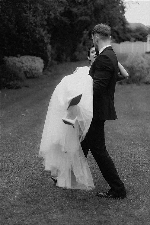 Wedding couple portrait of groom carrying bride in a tulle skirt, her heels lifted, on a garden lawn with trees and hedges behind