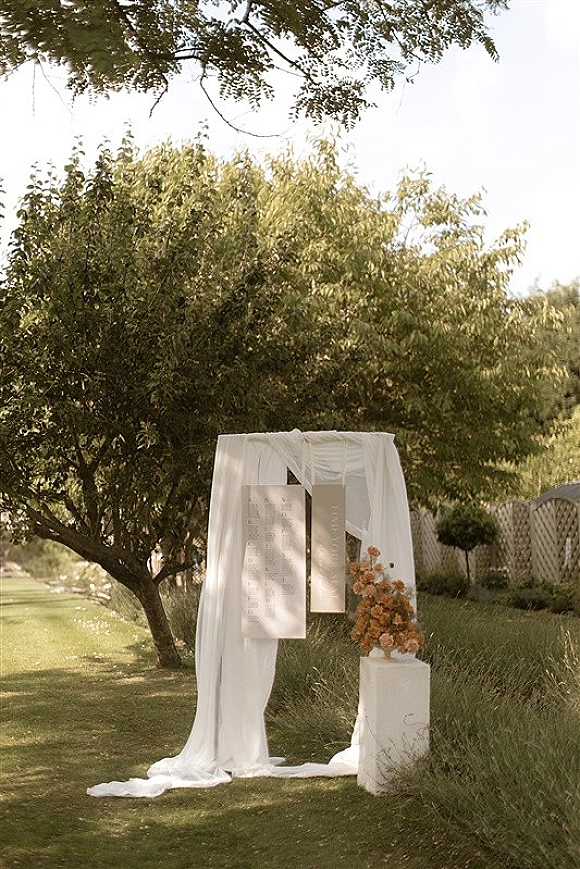 Ceremony backdrop with white draping fabric and hanging panels, plus a floral arrangement on a pedestal plinth on a garden lawn