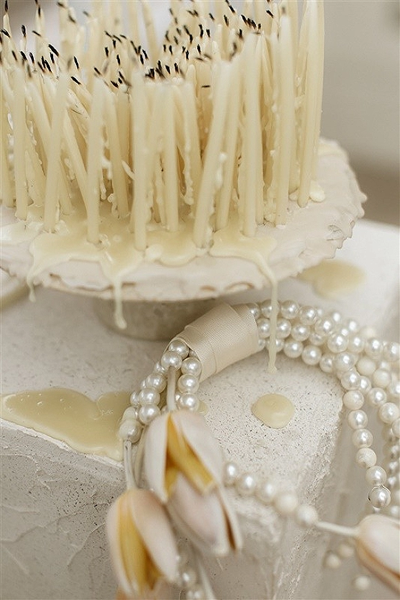 Wedding cake with white drip icing, textured buttercream, and a pearl necklace detail on a stand against a neutral wall