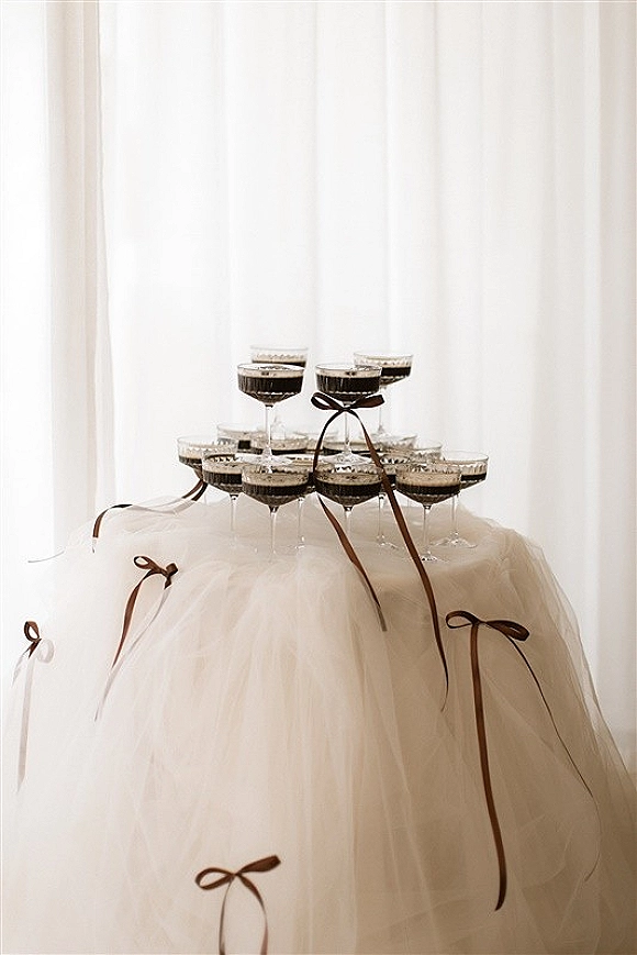 Champagne tower of coupe glass tower tiers filled with bubbly on a tulle-skirted table, accented by brown ribbon bows before white curtains