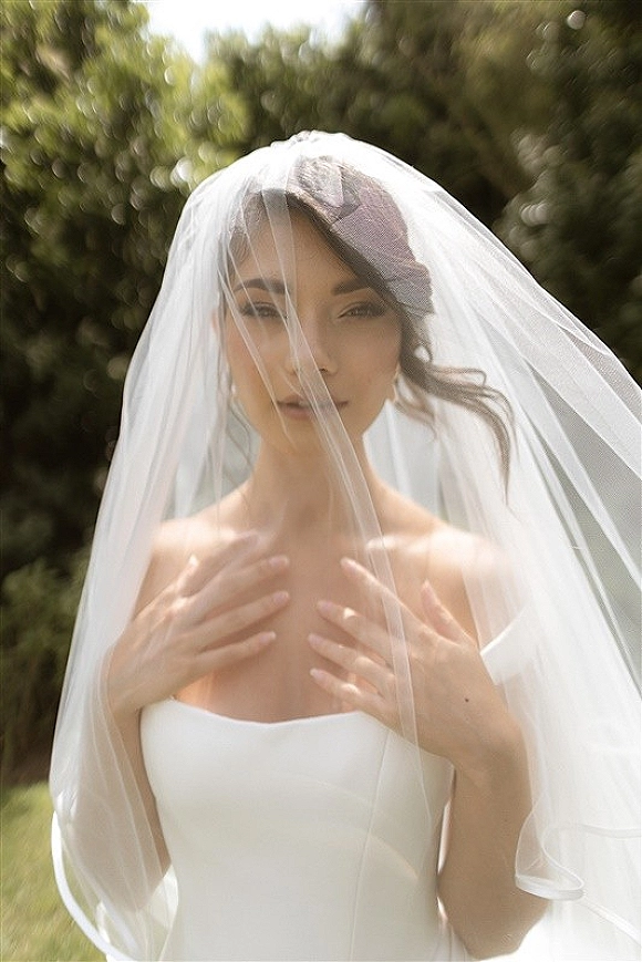Bridal portrait of a bride with veil draped over her face, wearing a strapless wedding dress and headpiece amid outdoor greenery
